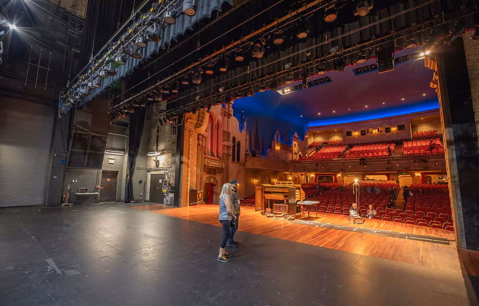 A view from offstage showing the transition between the dark backstage area and the bright wooden stage apron, where an organ sits facing the empty red theater seats under a blue-lit ceiling.