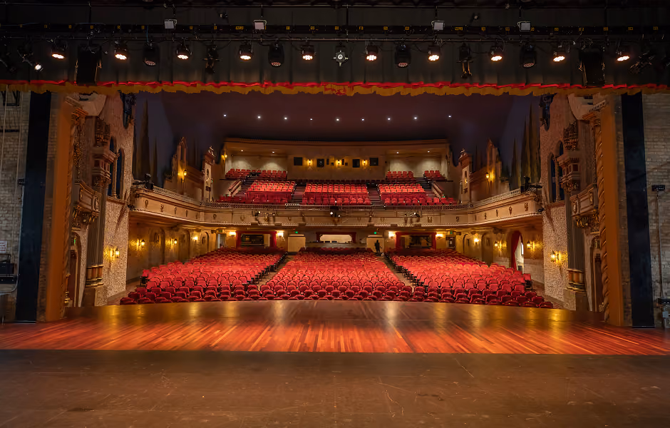 A view looking directly out from the center of the wooden stage into the vast, empty auditorium, highlighting rows of red seating on the main floor and upper balcony framed by stage lighting rigs above.