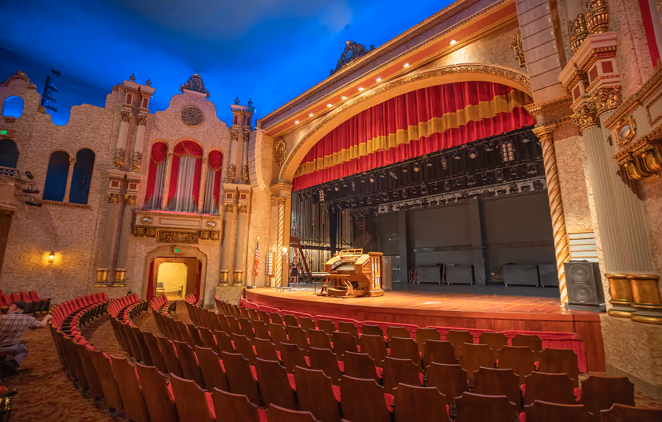 A low-angle view of a grand historic theater, showing rows of empty red seats facing a wooden stage framed by a large arch and red curtain. An vintage theatre organ sits stage right, next to elaborate plaster wall details under a deep blue painted sky ceiling.