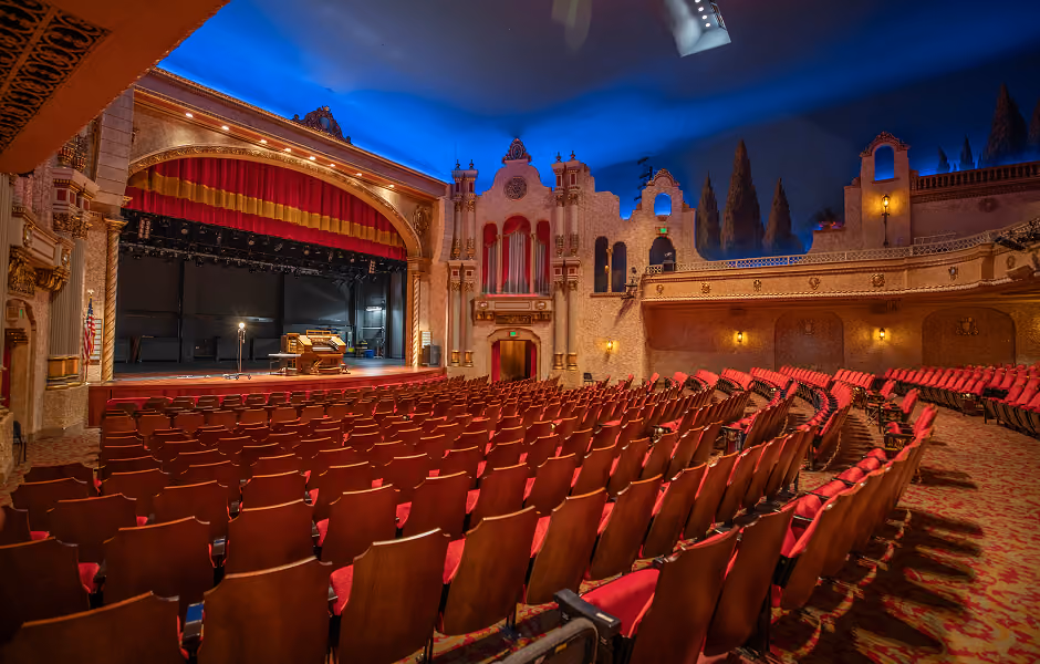 A wide shot from the main floor looking up toward the stage and upper seating level. The stage features a organ and lighting, set against Mediterranean-style architectural facades along the side walls and a vibrant atmospheric blue ceiling.