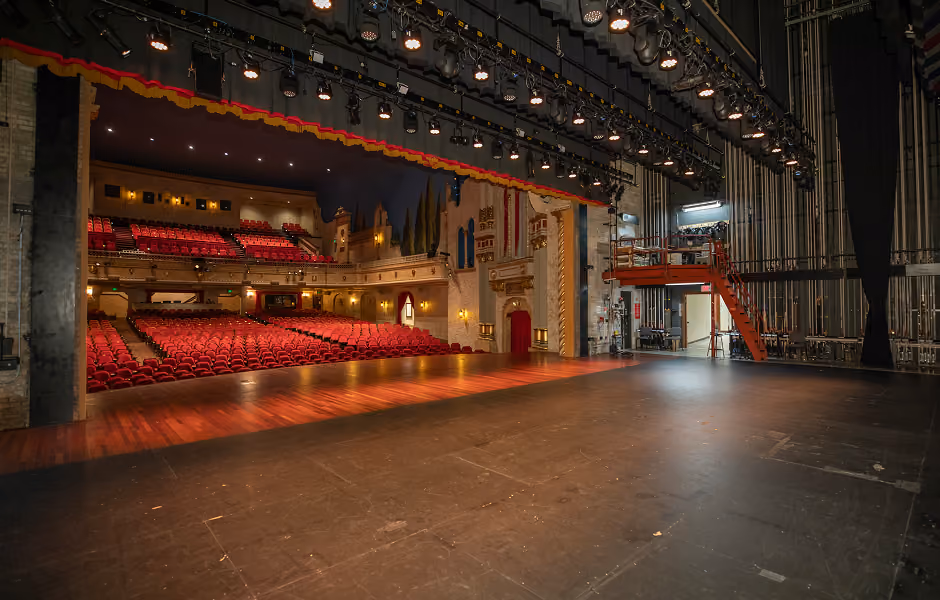 A wide shot from the side of the stage showing the expansive wooden stage apron, overhead lighting rigs, and offstage rigging infrastructure looking out into the empty auditorium seats.
