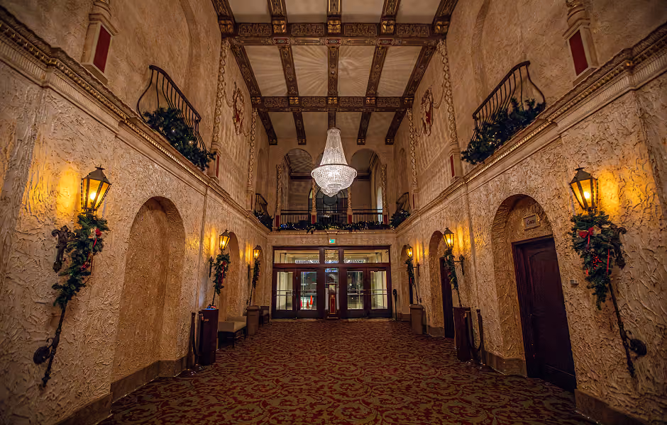 An eye-level view down a grand, grandly ornate theater lobby with textured tan walls, red patterned carpeting, and vintage lantern sconces adorned with holiday greenery. A large crystal chandelier hangs from a coffered ceiling, leading toward glass front doors in the background.