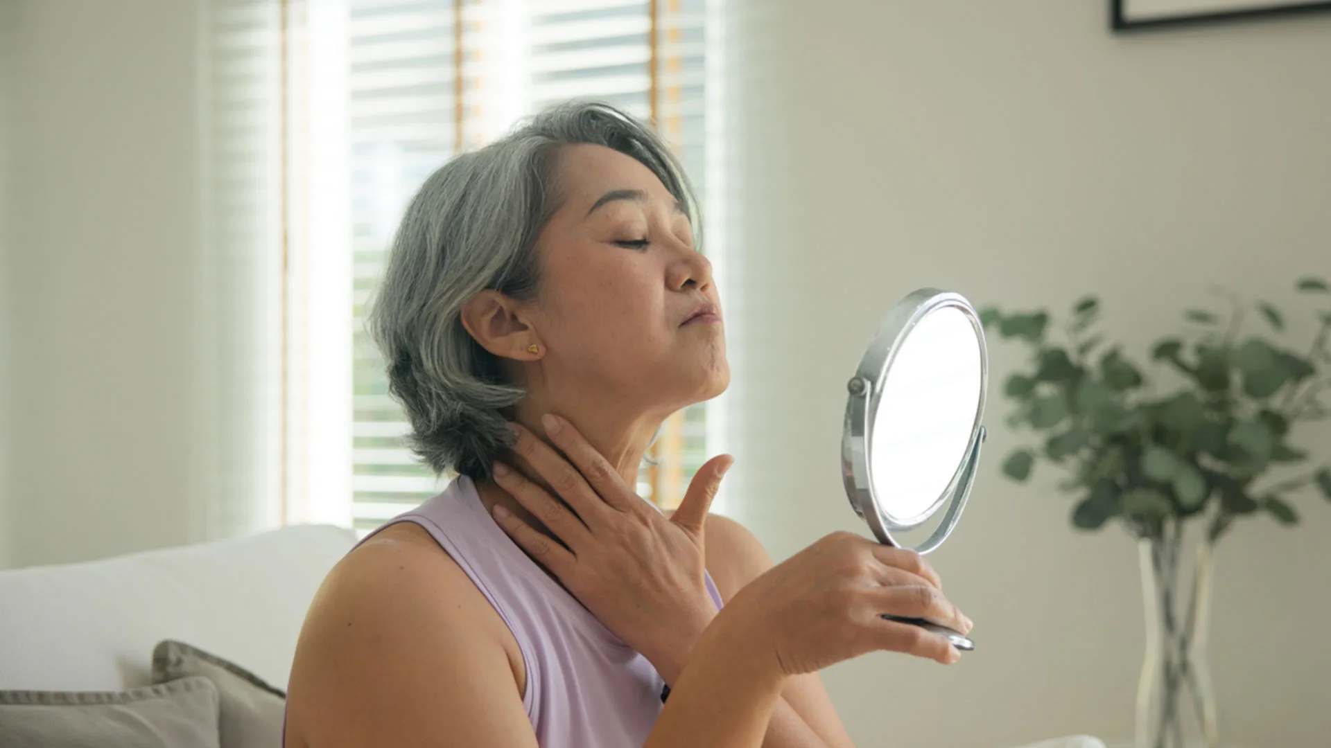 Mature woman checking her neck and jawline in a hand mirror at home