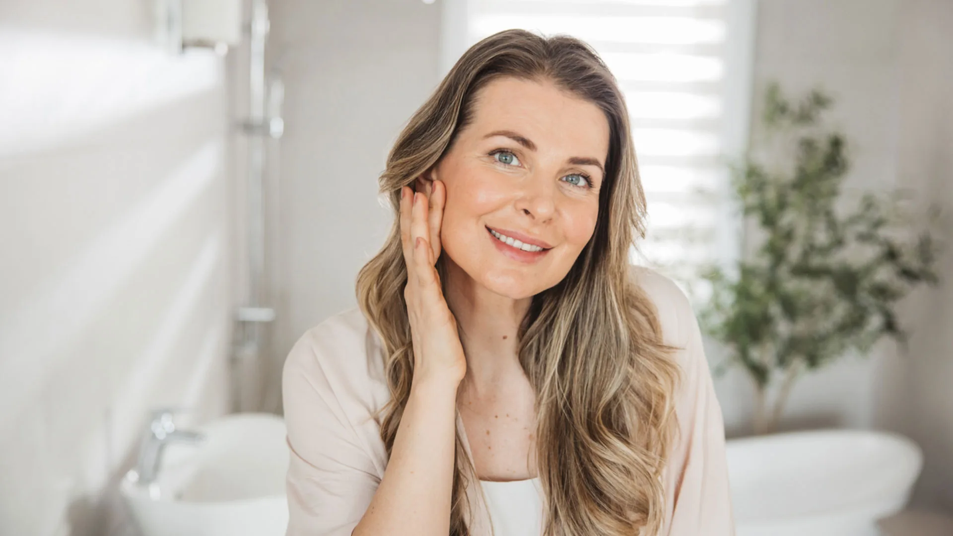 Smiling woman with long wavy hair touching her cheek in a bright bathroom