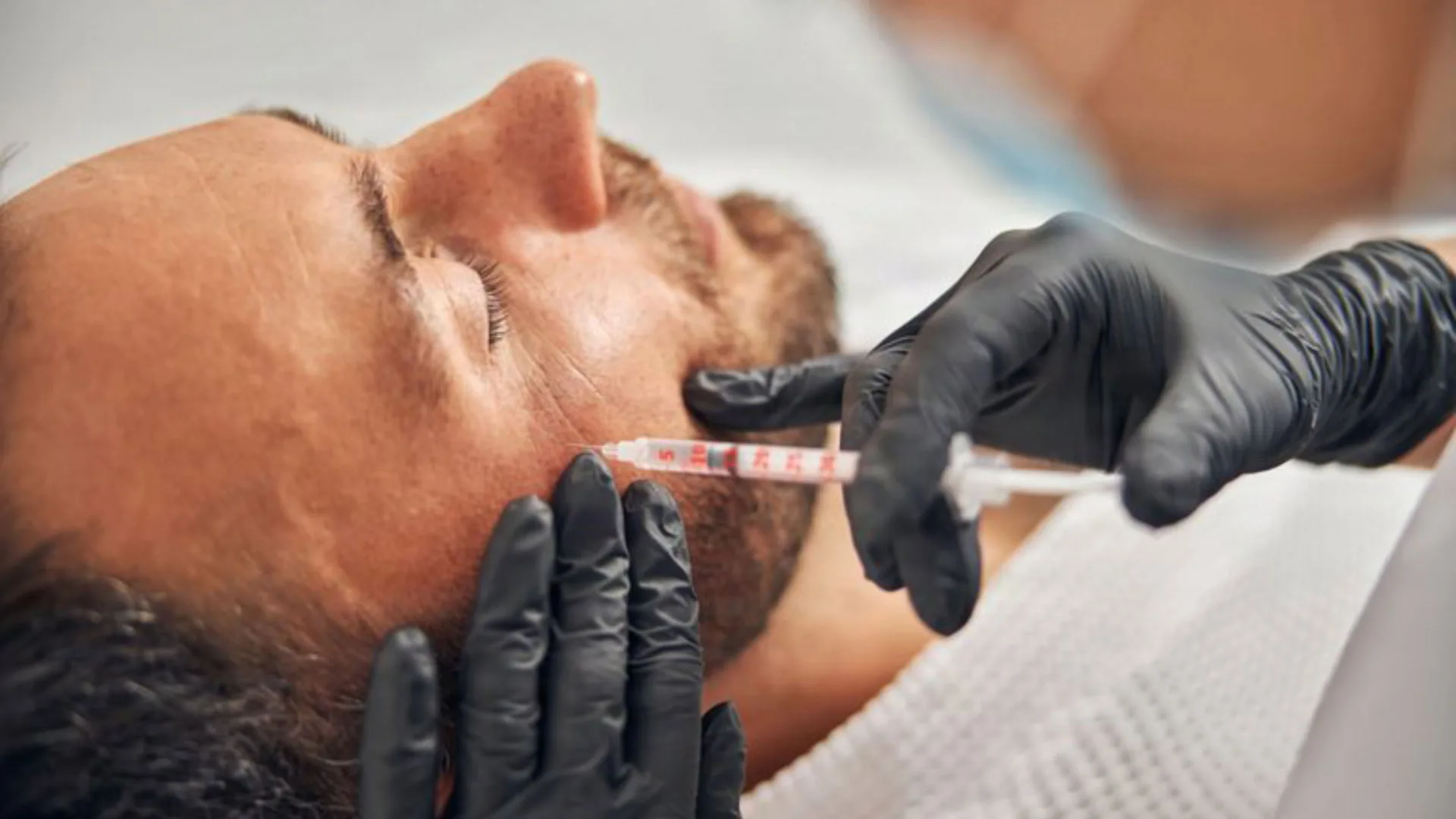 Gloved provider injecting a man's jaw area as he reclines with eyes closed