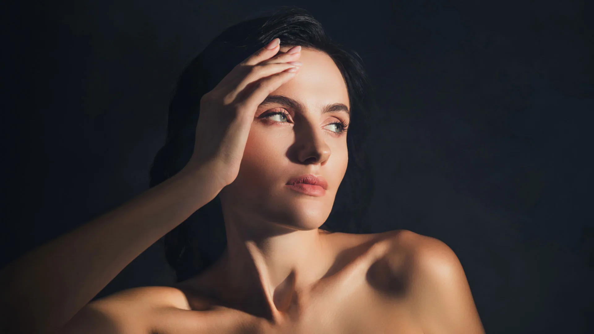 Woman with dark hair resting one hand on her forehead, softly lit against a dark background