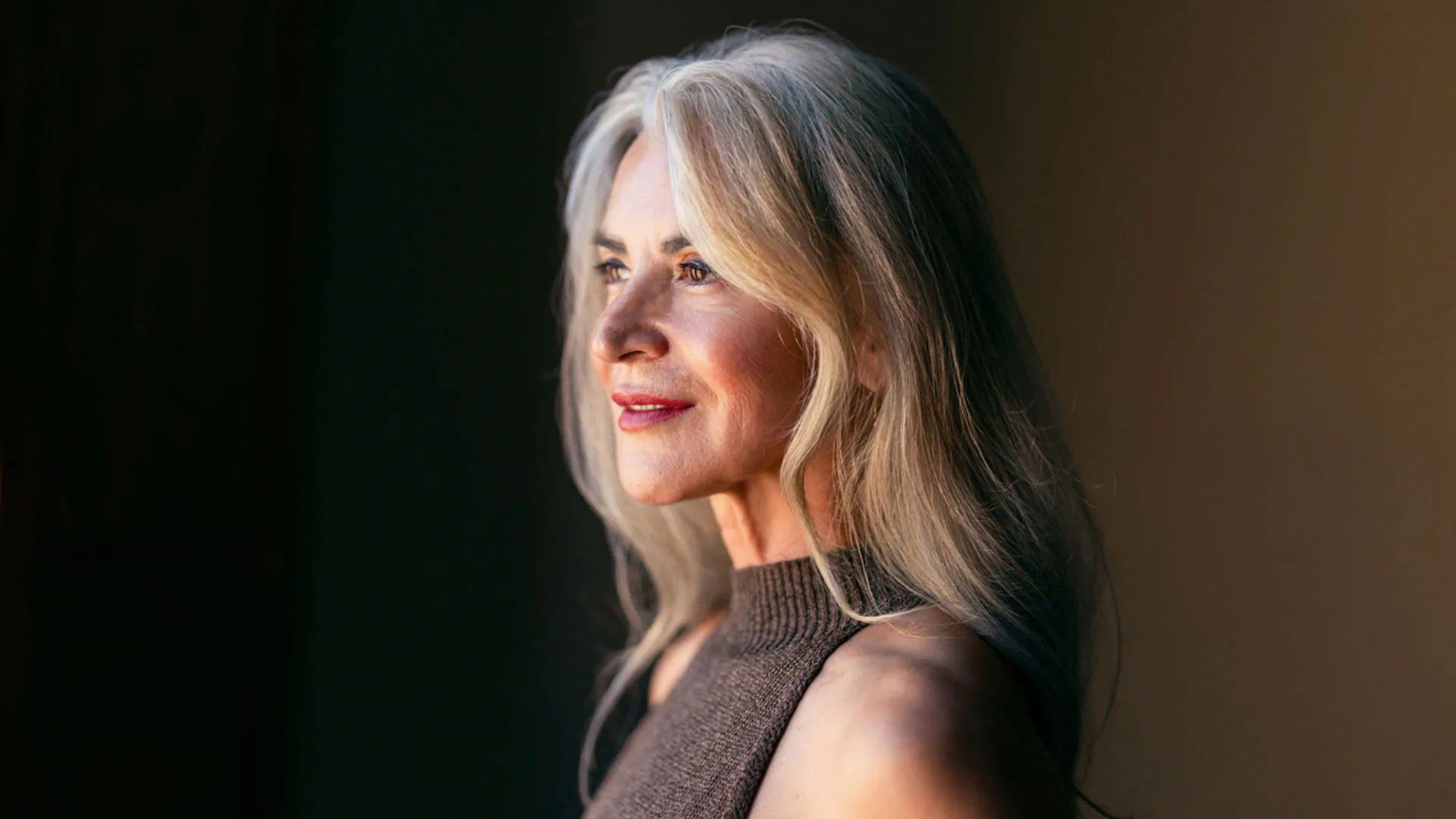 Mature woman with long silver hair gazing to the side in warm window light