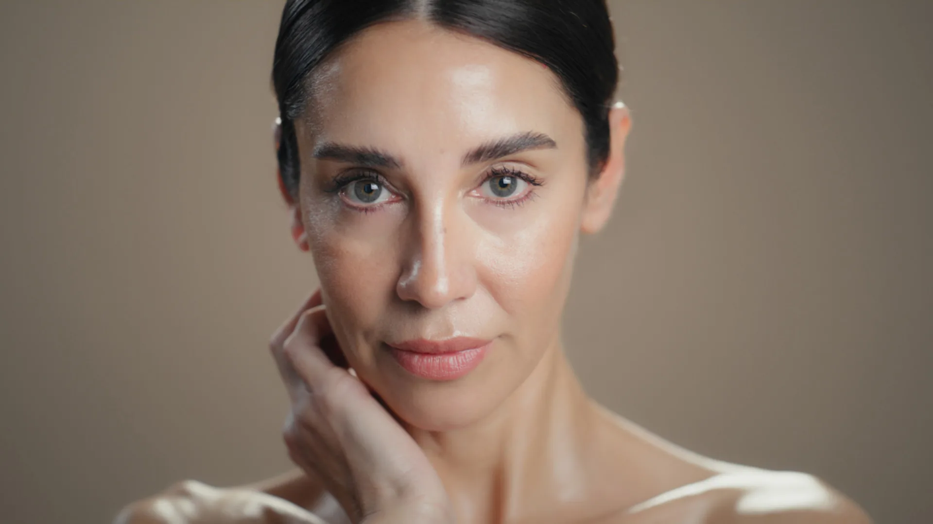Woman with dark hair pulled back touching her neck and looking at the camera