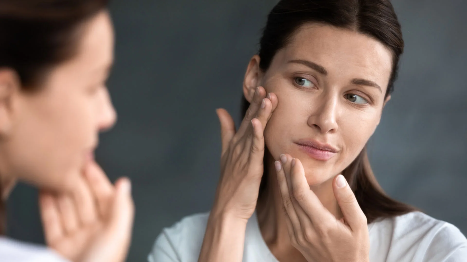 Woman examining her cheek with her fingertips while looking in a mirror