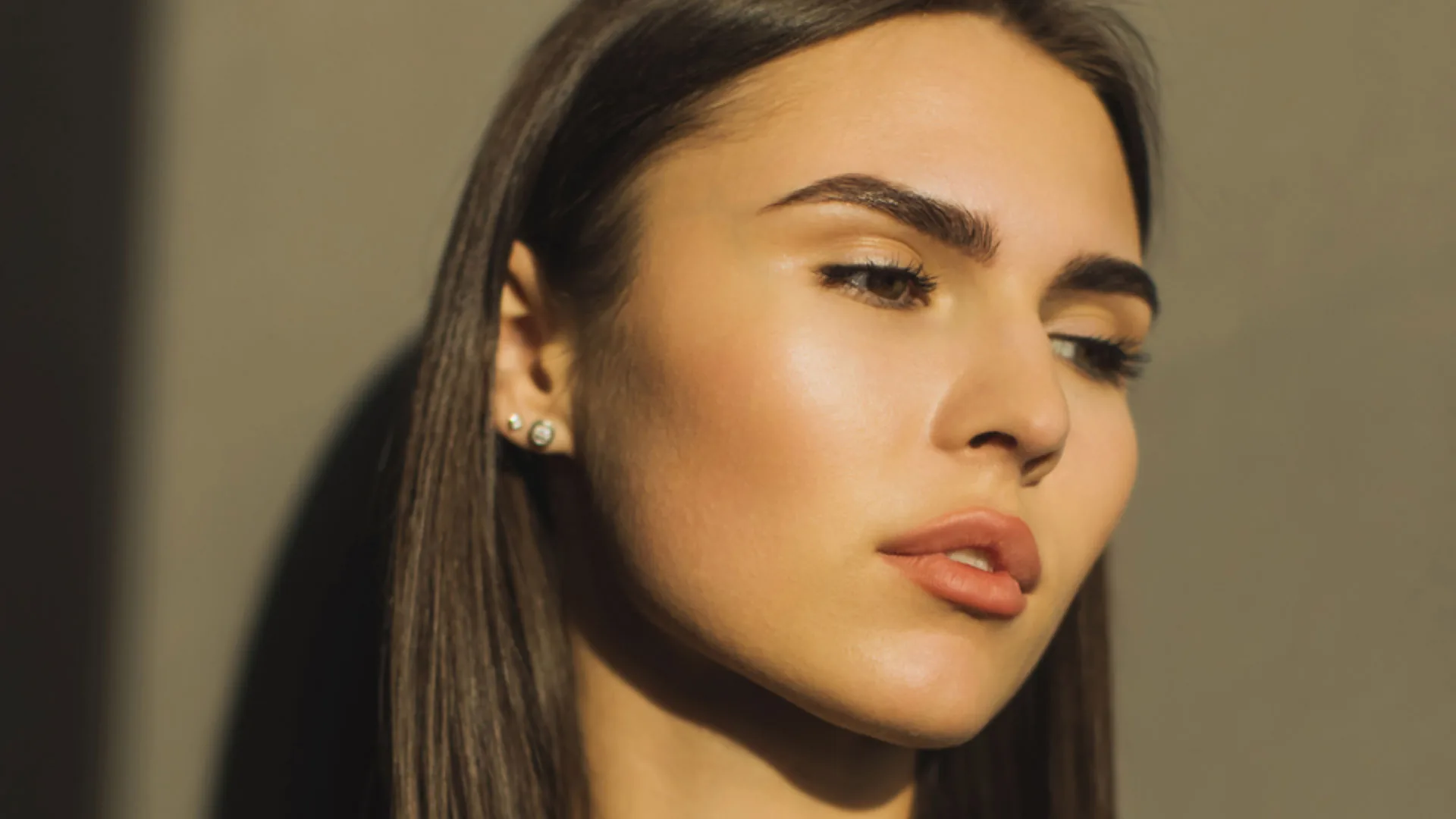 Woman with sleek dark hair and pearl earrings looking down in warm light