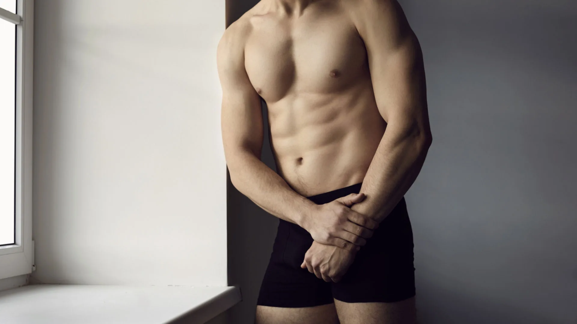 Shirtless man in black boxer briefs standing by a window, hands clasped in front
