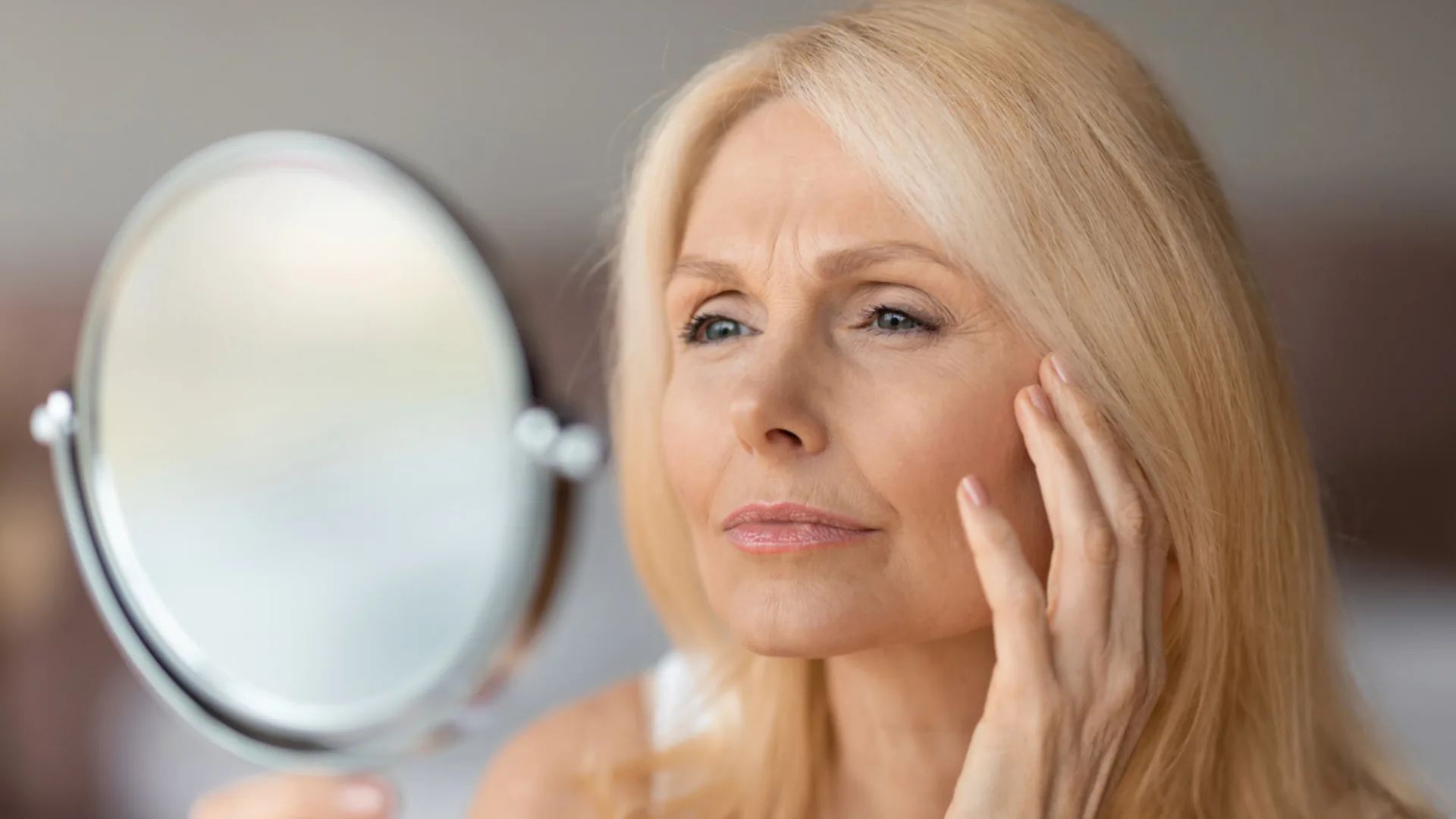 Mature woman with blonde hair touching her cheek while looking in a hand mirror