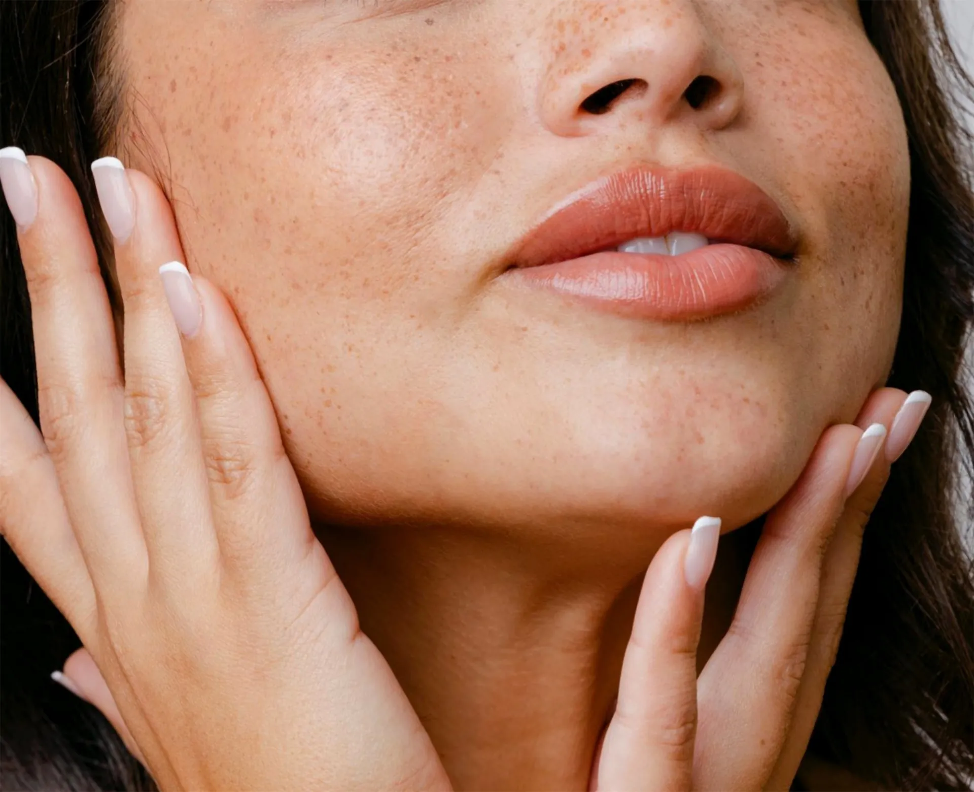 Close-up of a woman's freckled cheek, lips, and jaw, her fingertips framing her face