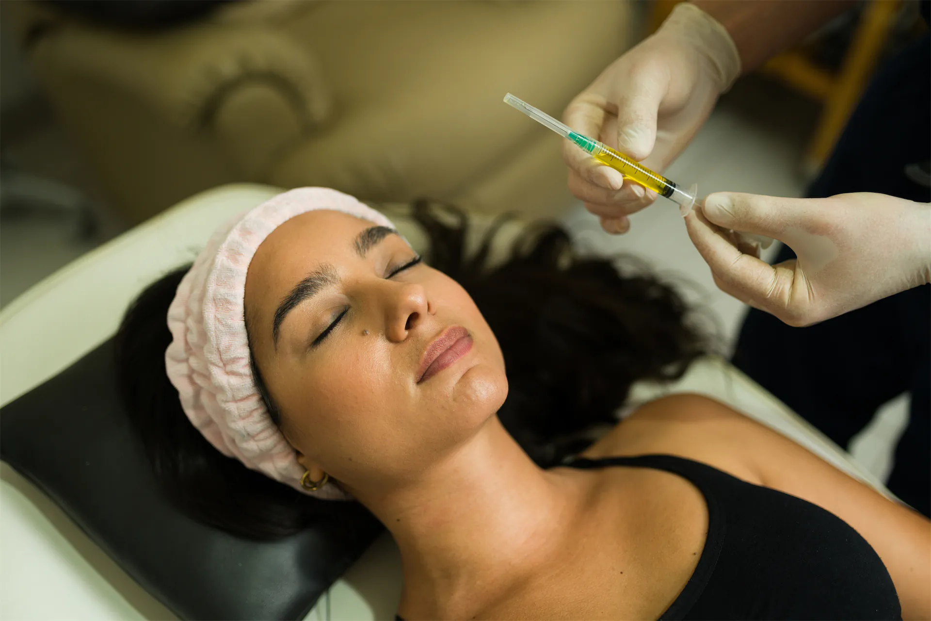 Gloved provider holding a syringe of golden fluid above a relaxed woman lying back in a headband