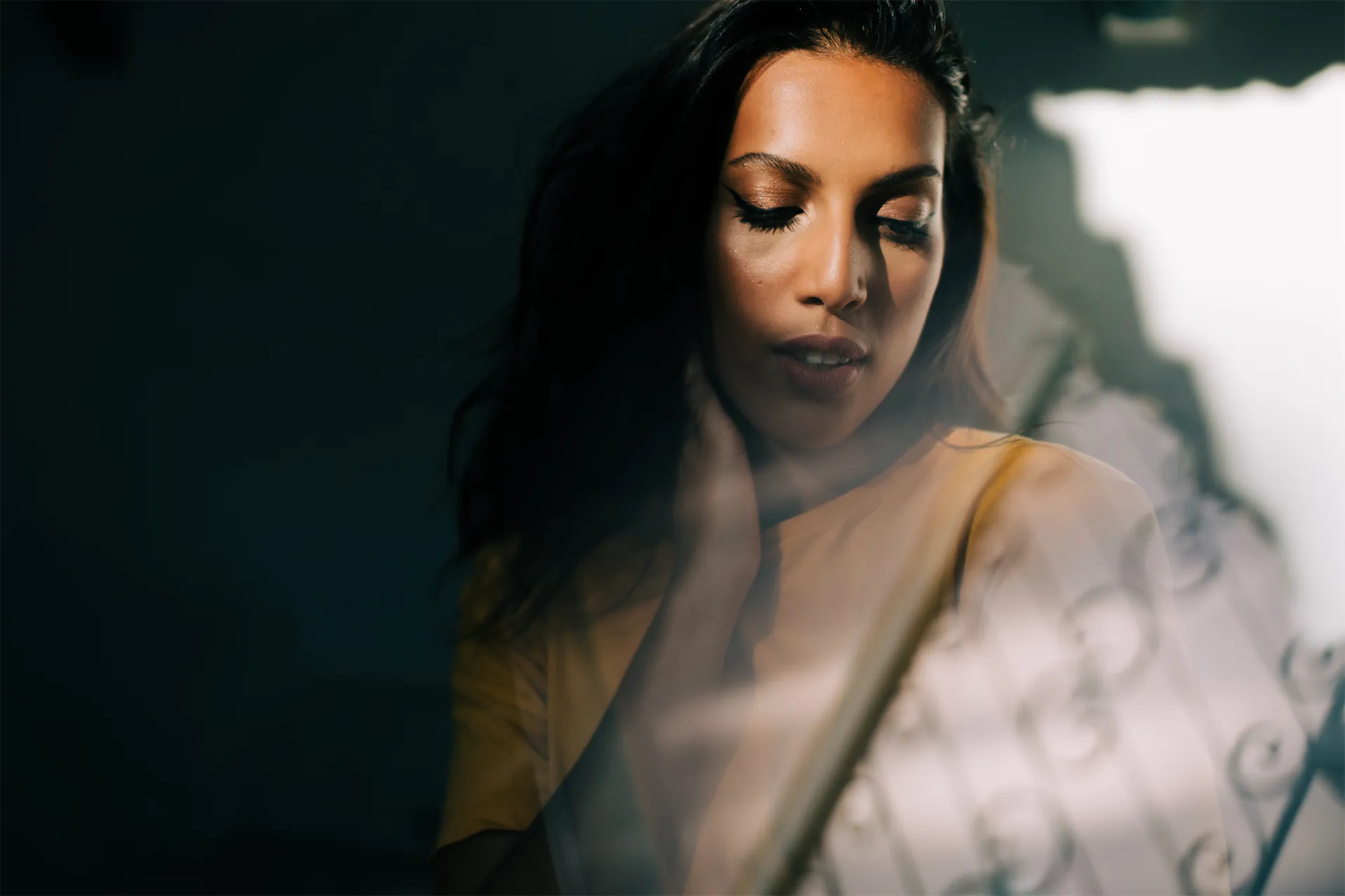 Woman with eyes lowered and a hand at her neck, lit by patterned light through a window