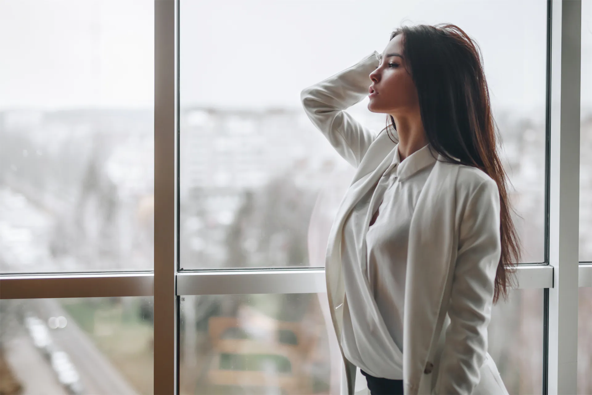 Woman with long dark hair in a white blazer gazing out a tall window