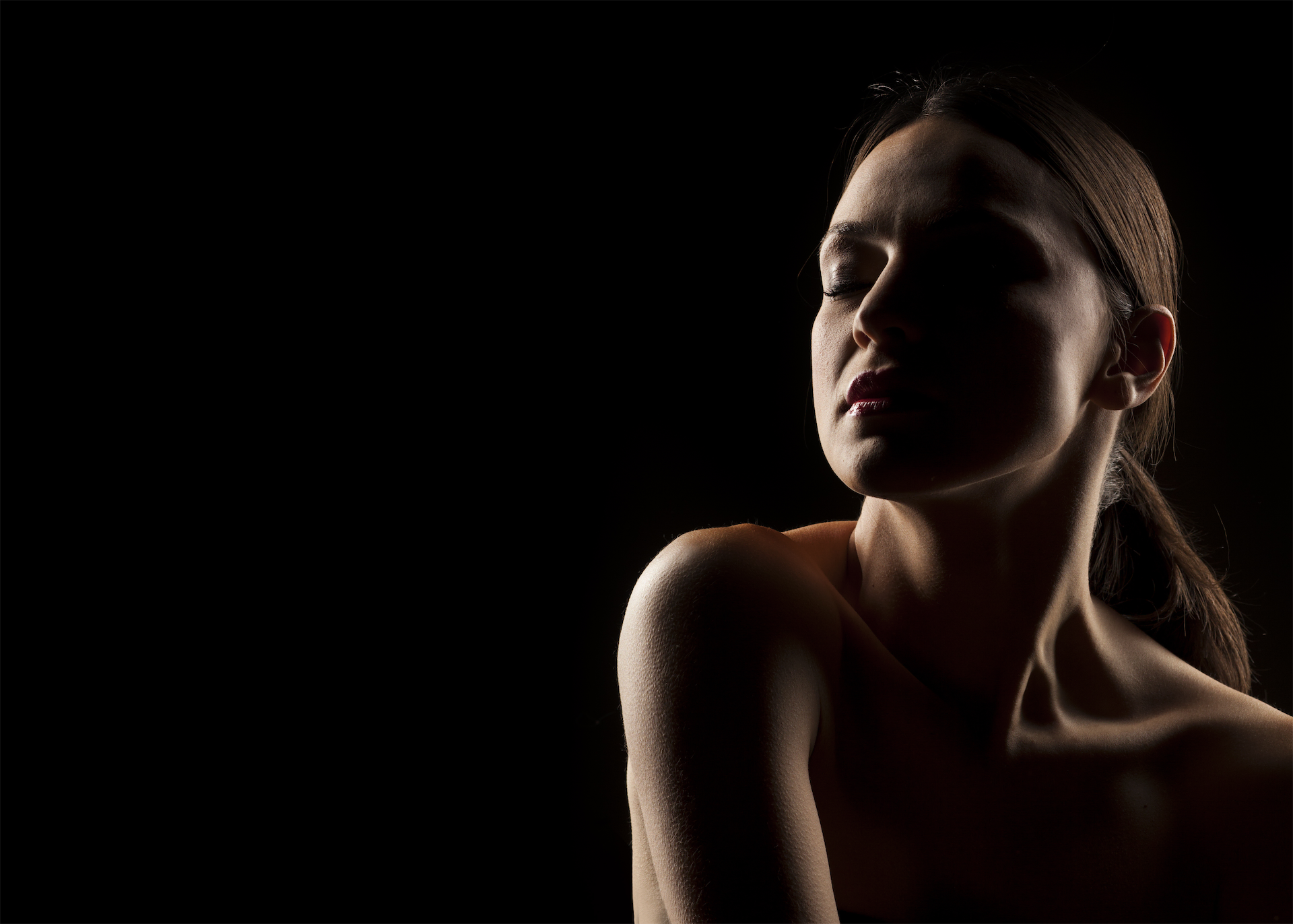 Woman with eyes closed and her hair pulled back, side-lit against a black background