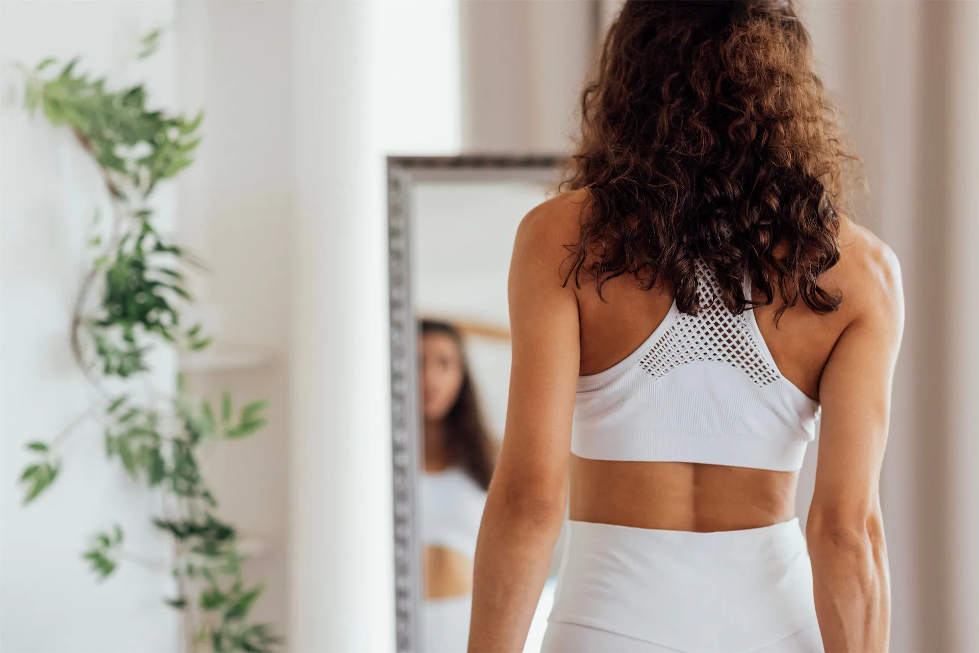 Woman in white activewear seen from behind, looking at her reflection in a mirror
