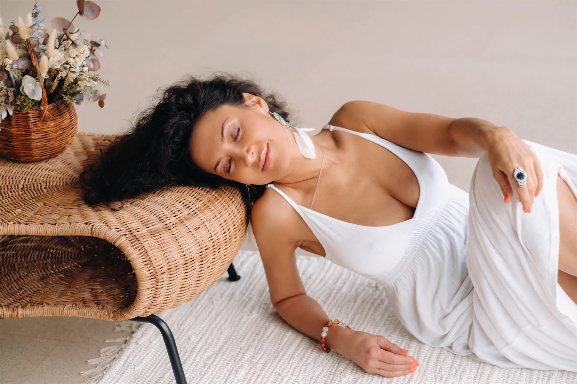 Smiling woman in a white slip dress reclining with eyes closed beside a wicker chair