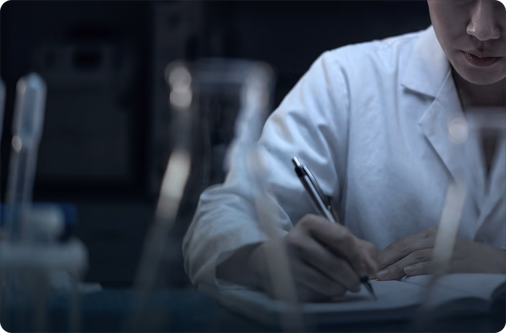 Scientist in a lab coat writing notes with lab glassware in the foreground.