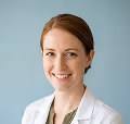 Smiling woman with red hair wearing a white lab coat against a light blue background.