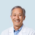 Smiling elderly Asian man with gray hair wearing a white coat and blue shirt.