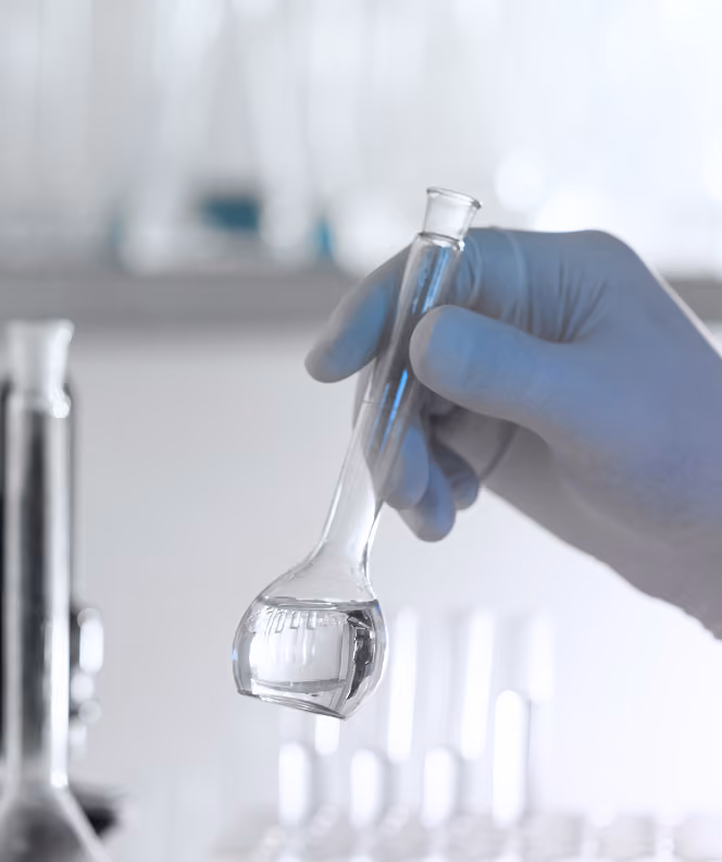 Gloved hand holding a small clear glass flask with liquid in a laboratory setting.