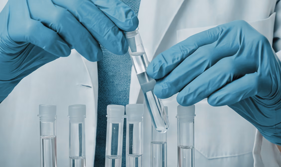 Hands in blue gloves holding a test tube and dropper over multiple test tubes with clear liquid.