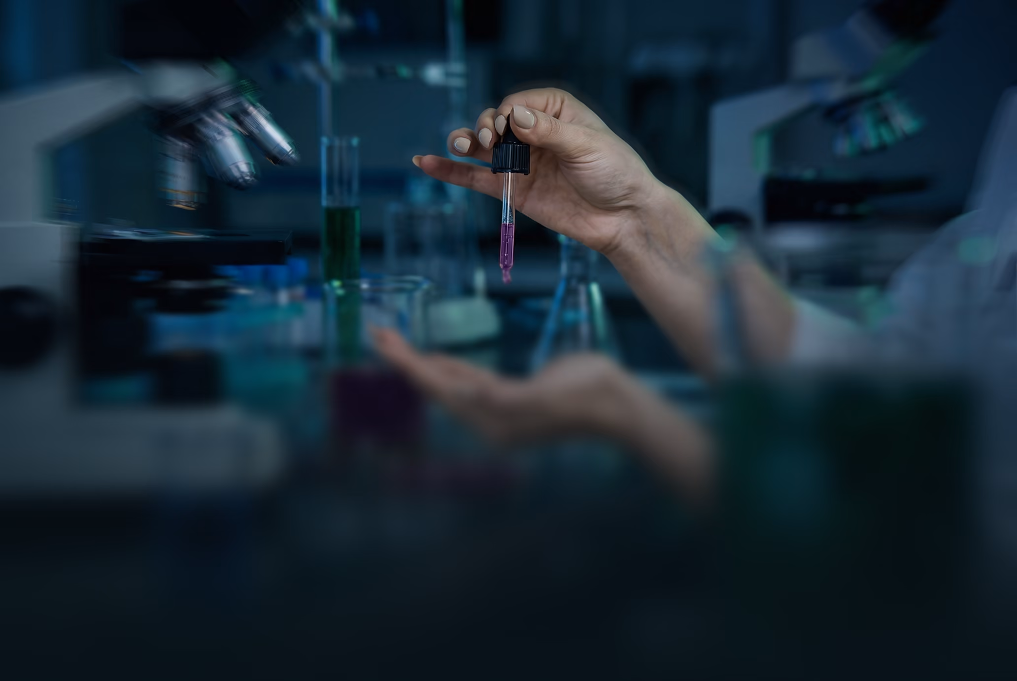 Scientist's hand holding a dropper with purple liquid over lab glassware in a dim laboratory.