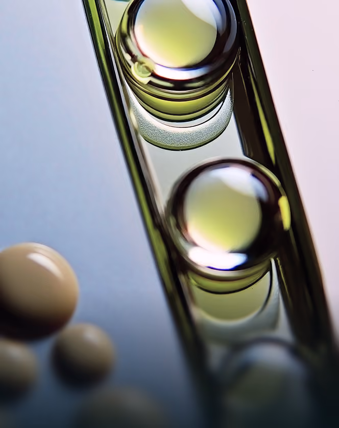 Close-up of transparent glass vial with greenish-yellow reflective liquid inside and blurred pills nearby.