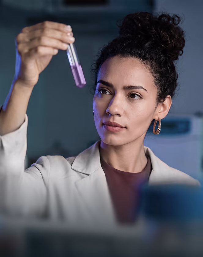 Scientist in a lab coat holding and examining a test tube with purple liquid.