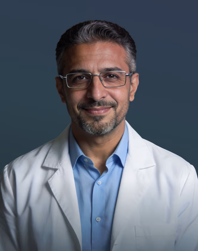 Smiling middle-aged male doctor with glasses, gray hair, blue shirt, and white coat against dark background.