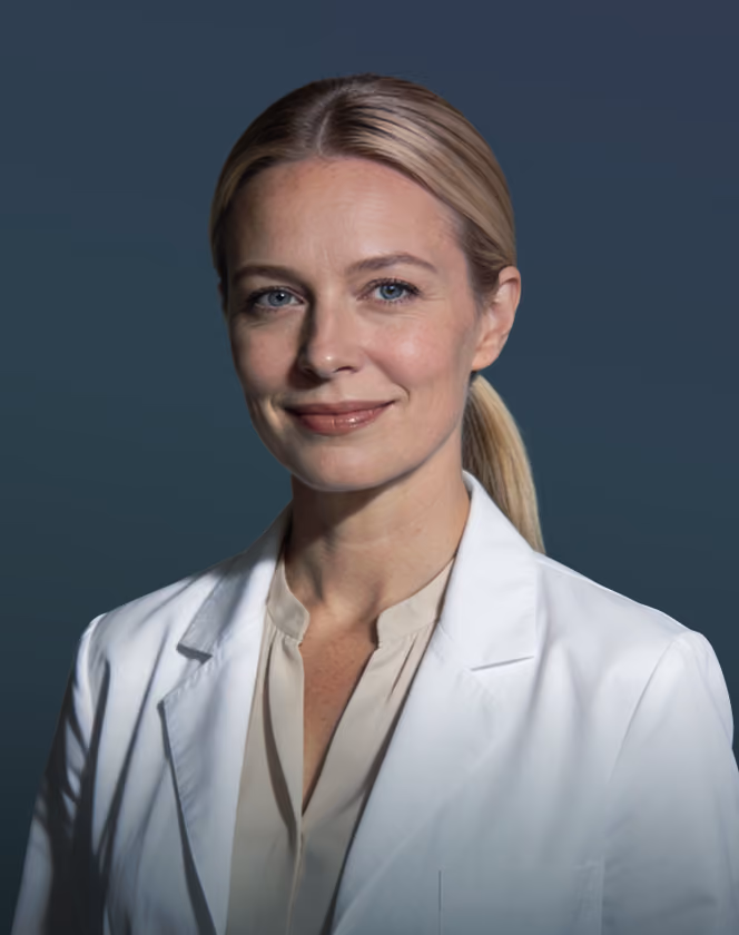 Smiling female doctor with blonde hair in a white coat and beige blouse against a dark background.