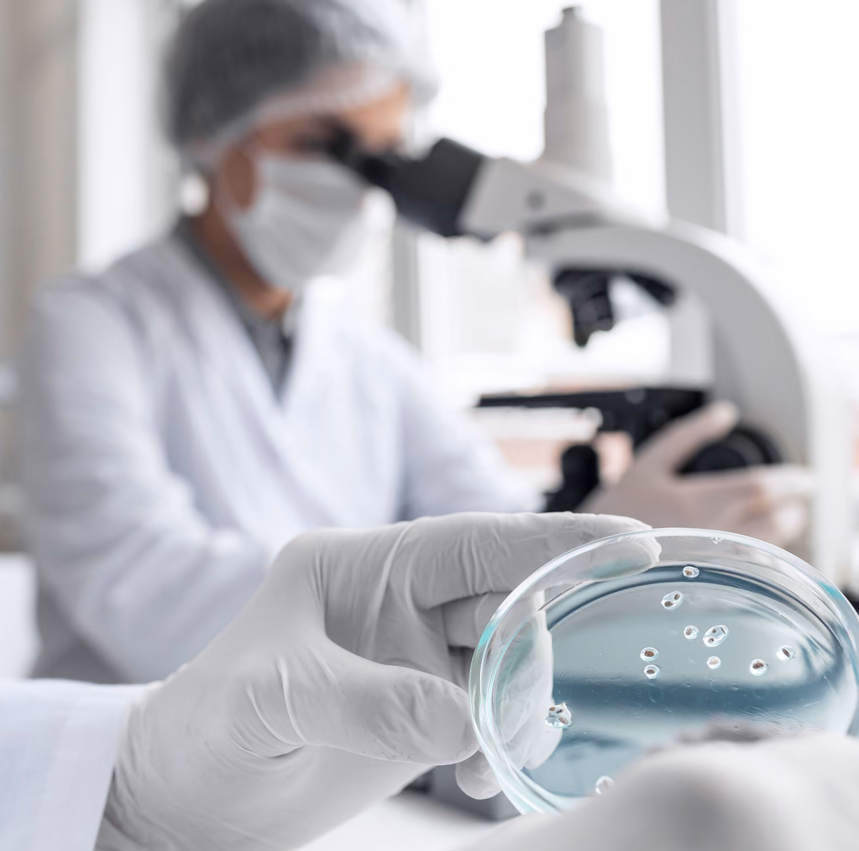 Scientist in protective gear holding a petri dish with samples, microscope in background.