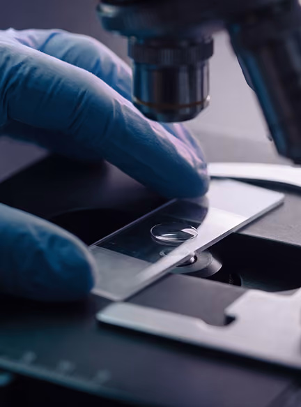Gloved hand placing a glass slide with liquid under a microscope lens.