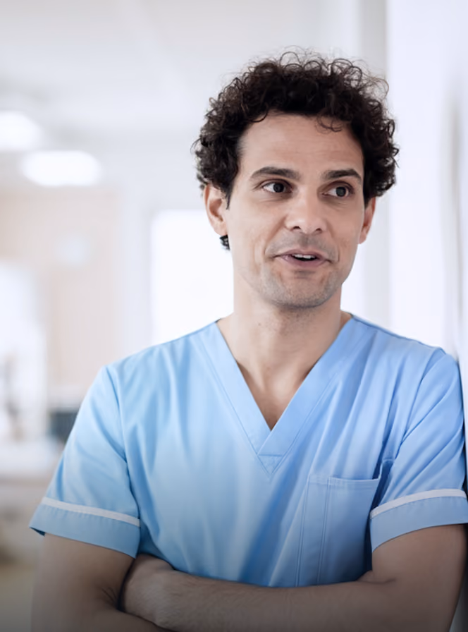 Male healthcare worker in light blue scrubs standing with arms crossed in a bright clinical setting.