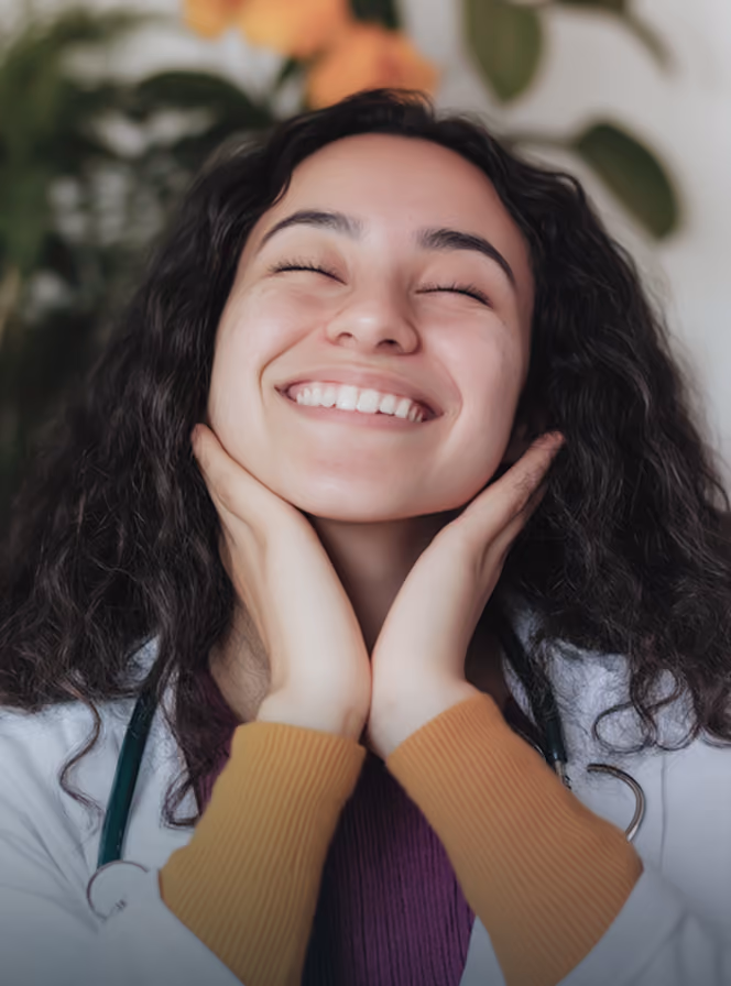 Smiling woman with curly hair wearing a white coat and stethoscope, holding her face with both hands.