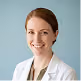 Smiling woman with brown hair wearing a white lab coat against a blue background.