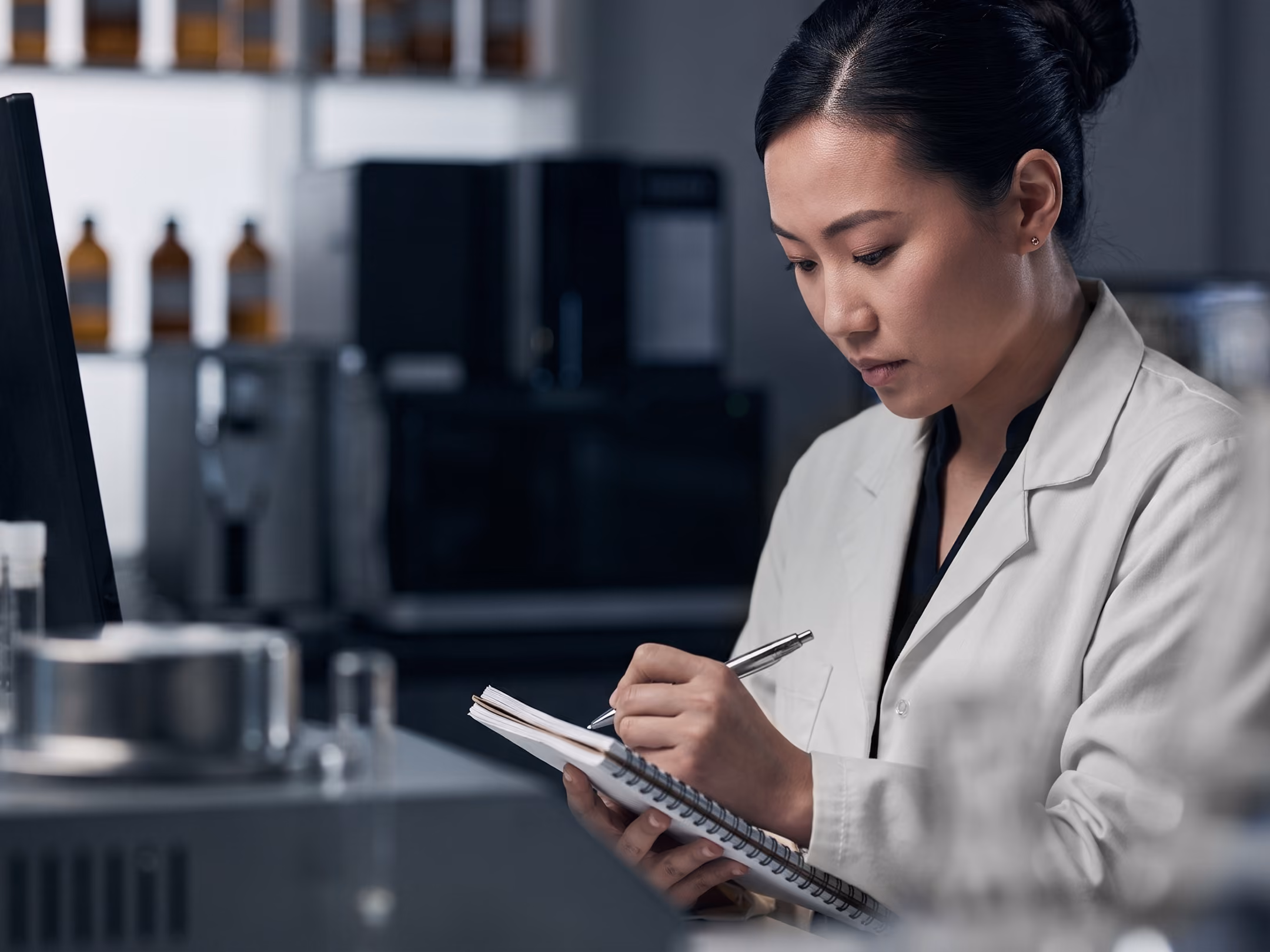 Scientist in a lab coat writing notes in a notebook inside a laboratory setting.