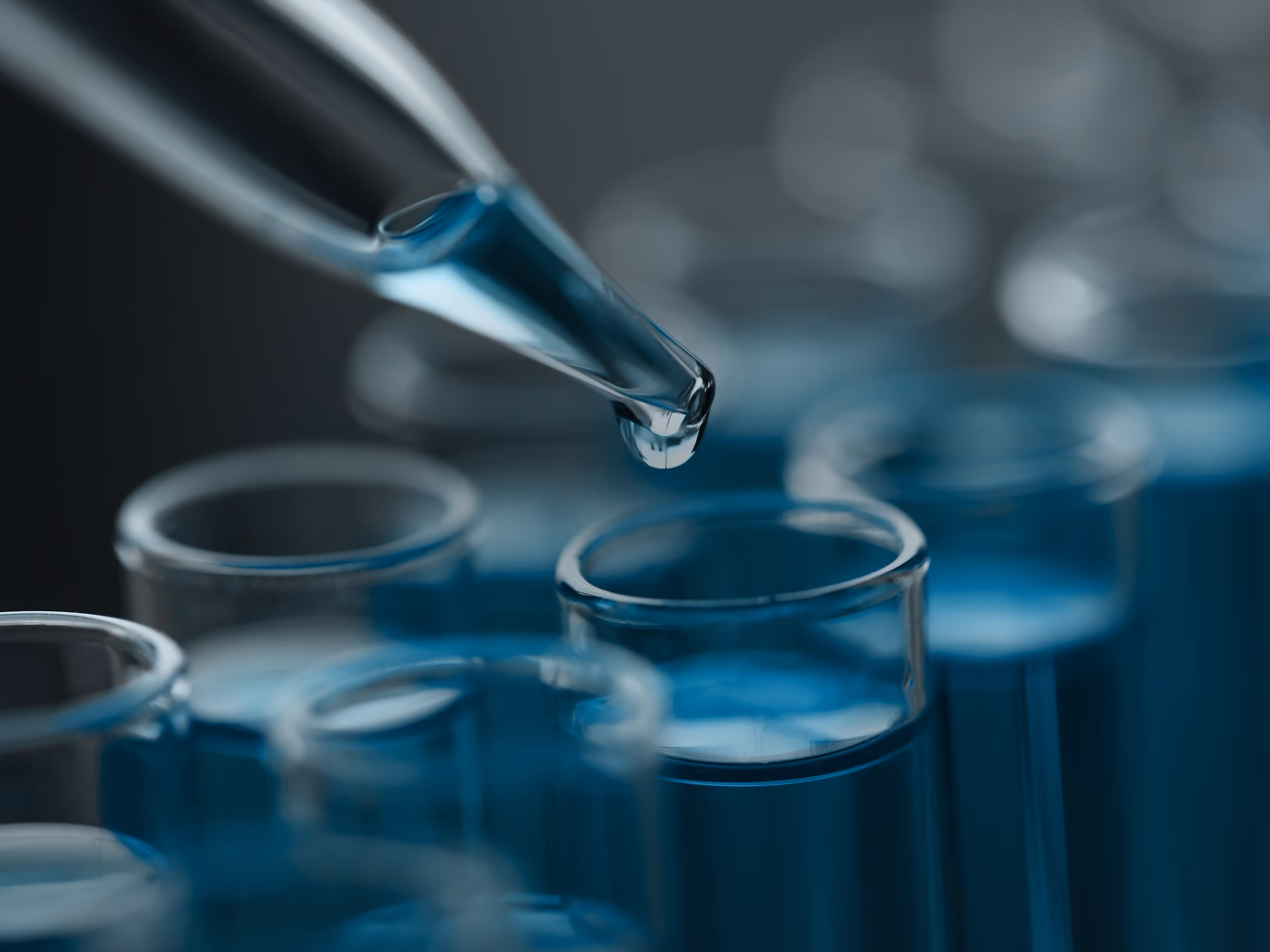 Pipette dropping blue liquid into a glass test tube among other tubes filled with blue liquid.