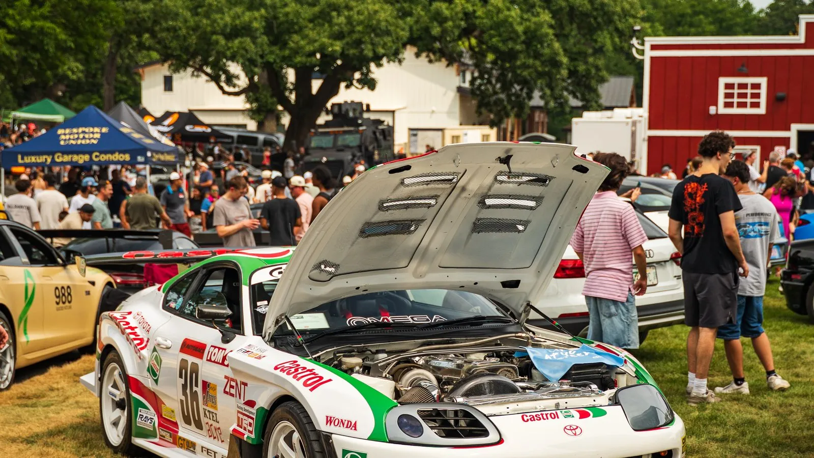 A liveried race car with its hood raised, drawing a crowd beside the red barn