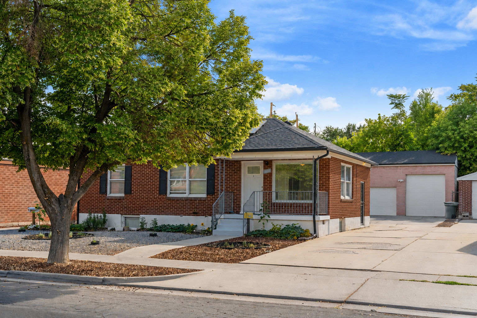 Single-story brick house with a large front tree, concrete driveway, and detached garage under blue sky.