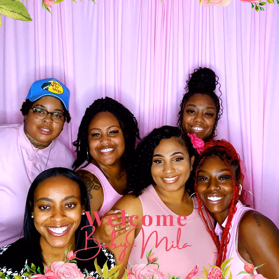 Six smiling women posing in front of a pink curtain with floral decorations.