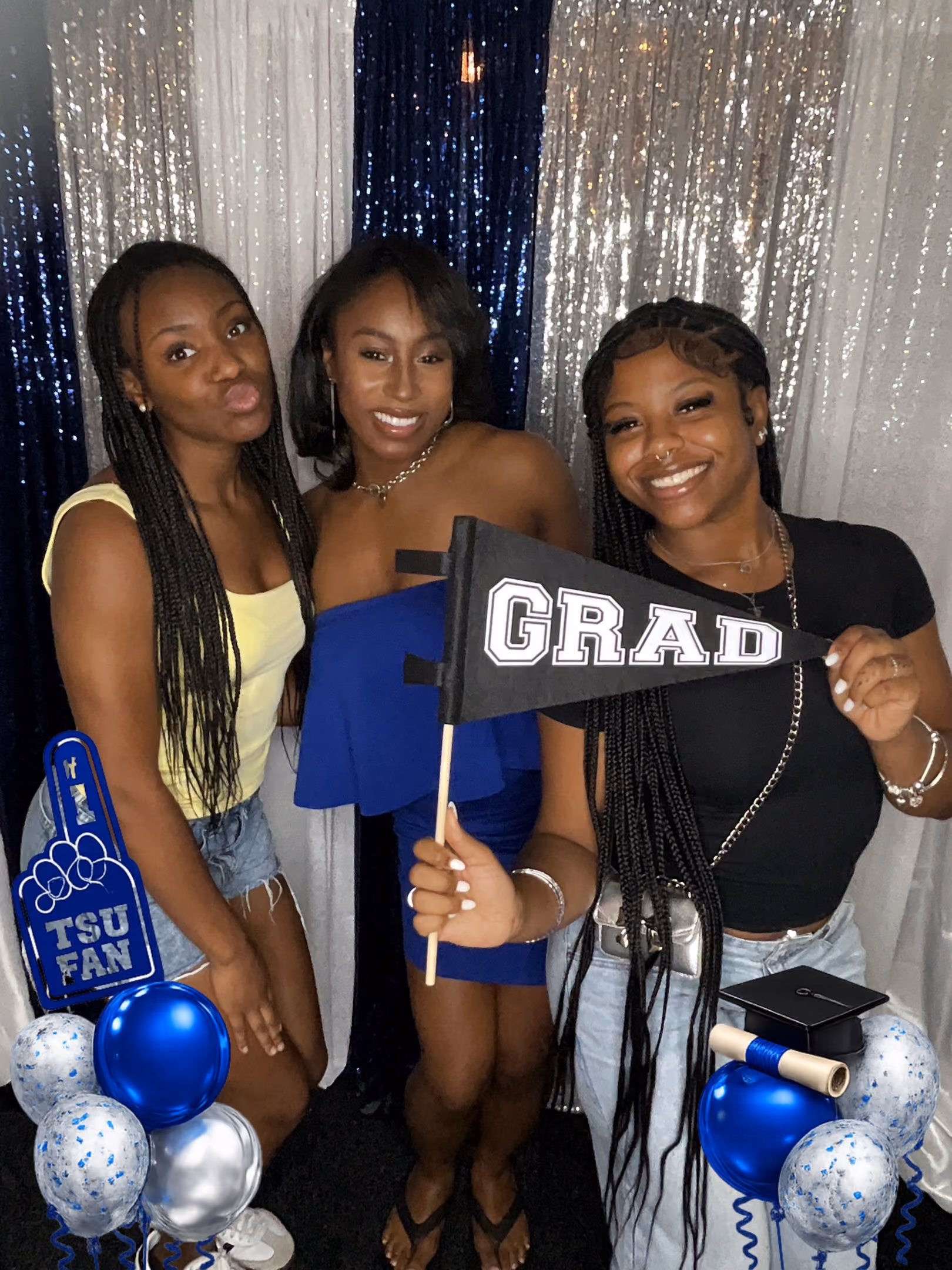 Three women smiling in front of blue and silver curtains at a graduation celebration with balloons and a grad pennant.