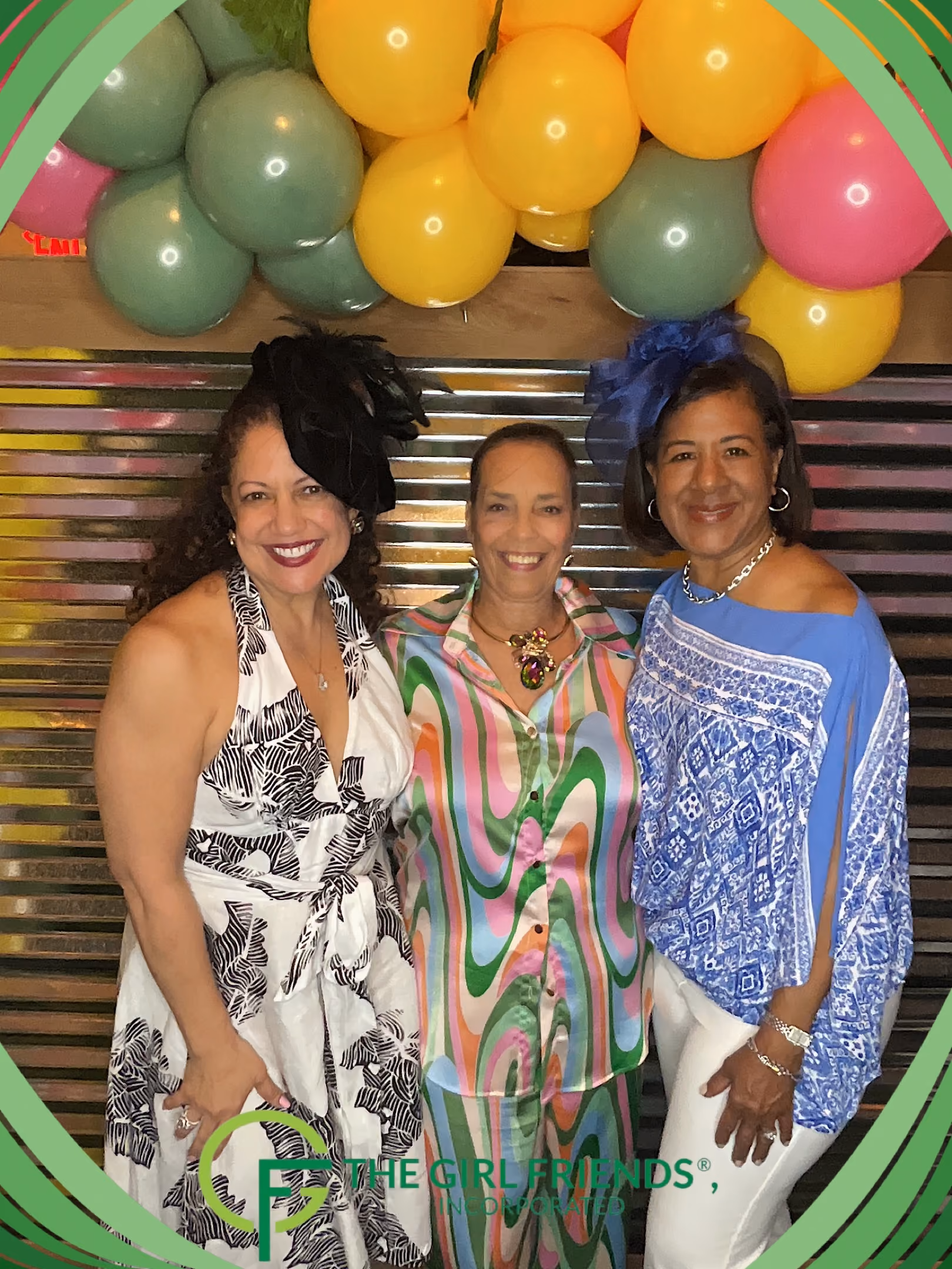 Three smiling women posing in colorful attire under a balloon arch with green frame.