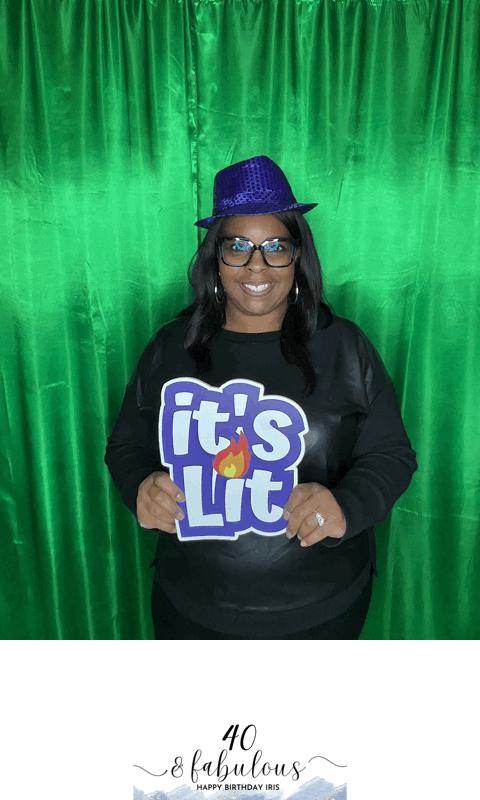 Smiling woman in glasses and purple hat holding a sign that says "it's Lit" against a green curtain.