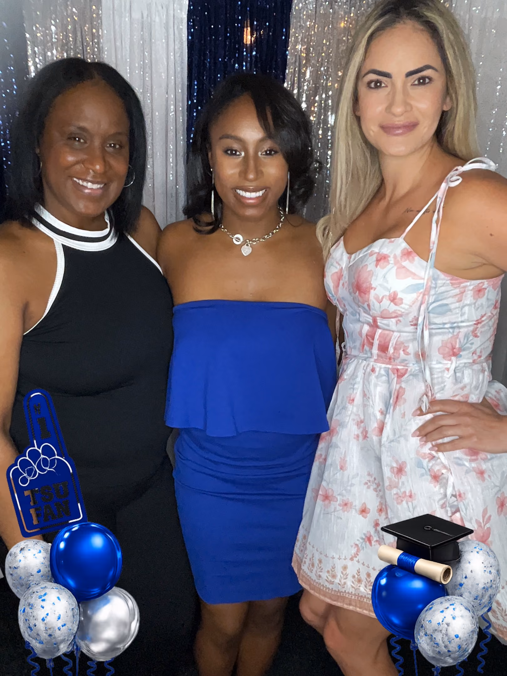 Three women smiling in front of a shiny backdrop with graduation-themed balloon decorations.