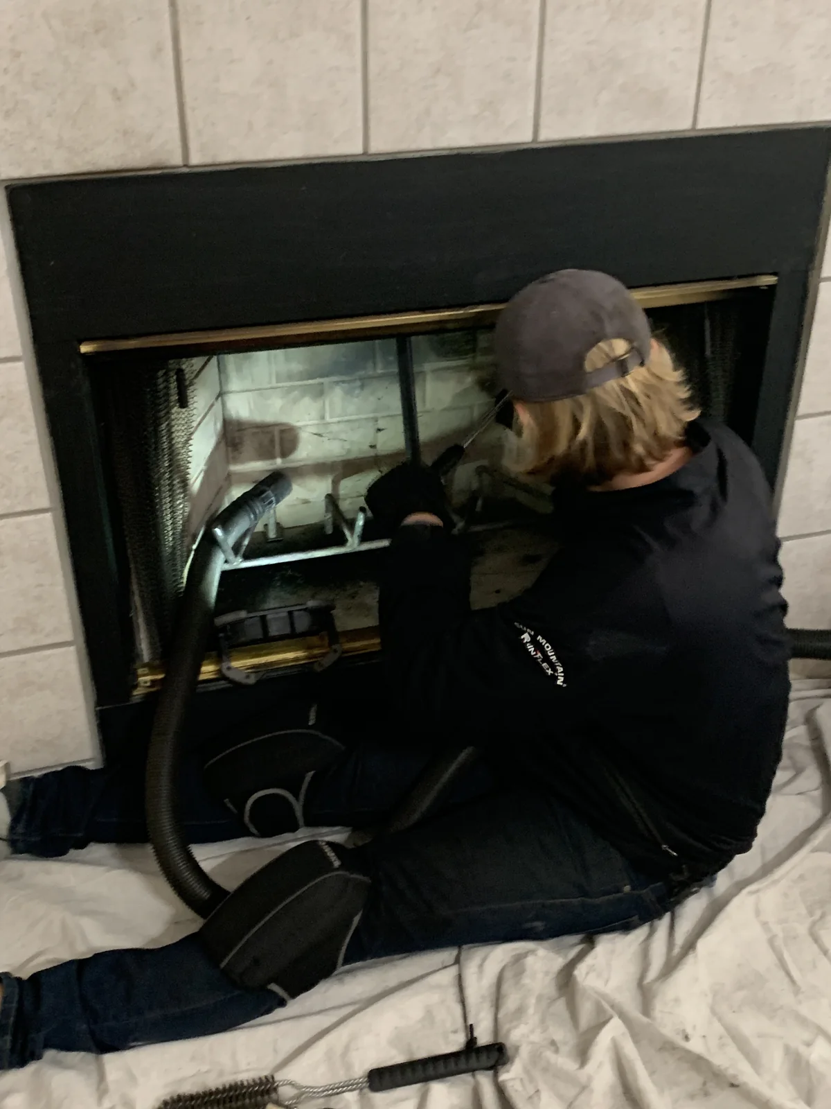 A chimney sweep technician kneeling at an open fireplace with protective covering laid over the hearth and floor.