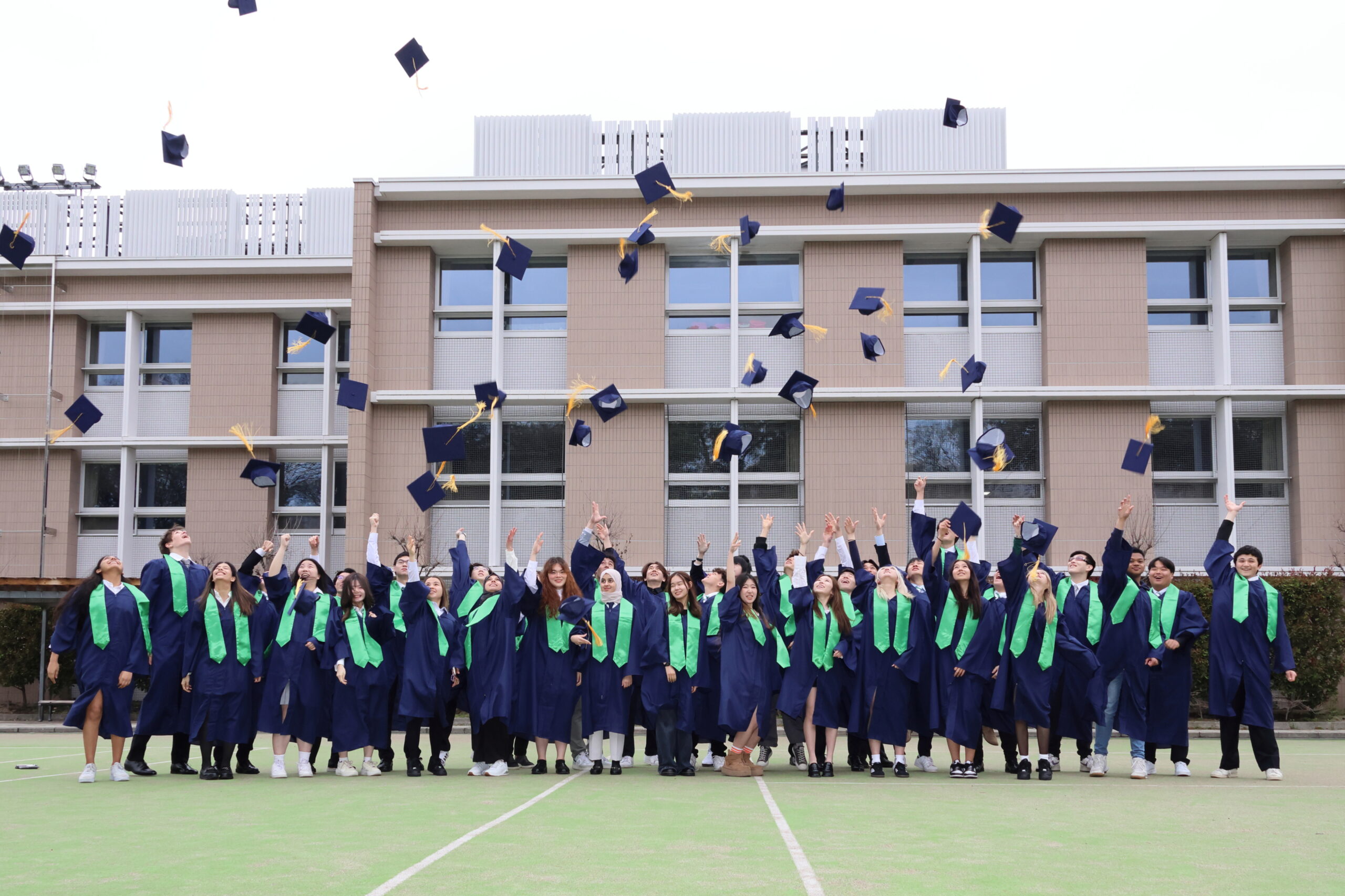 Aoba-Japan International School high school graduates wearing graduation robes and throwing their graduation caps in celebration while standing outdoors on a school field