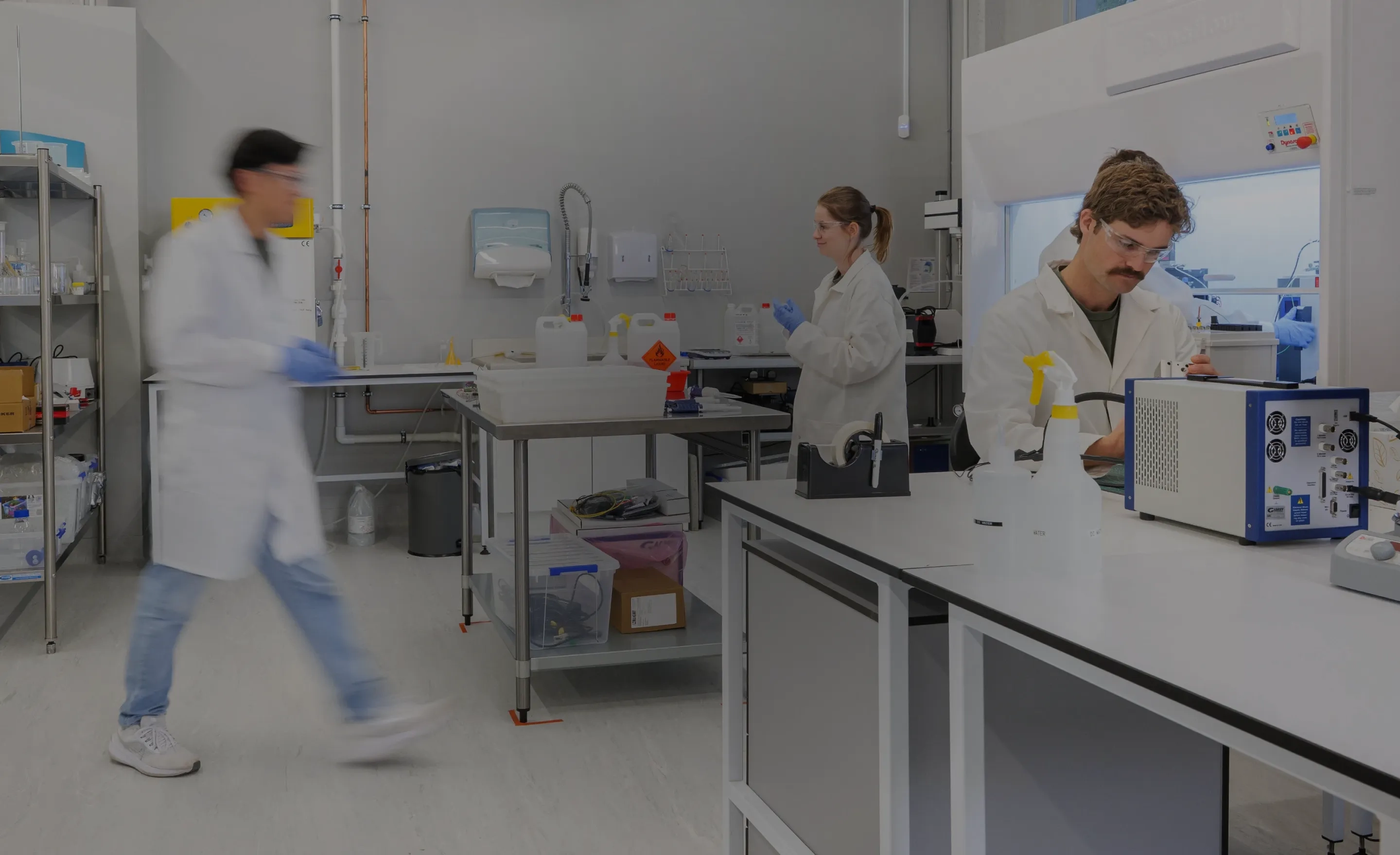 Three scientists in lab coats working in a laboratory with equipment and chemical containers.